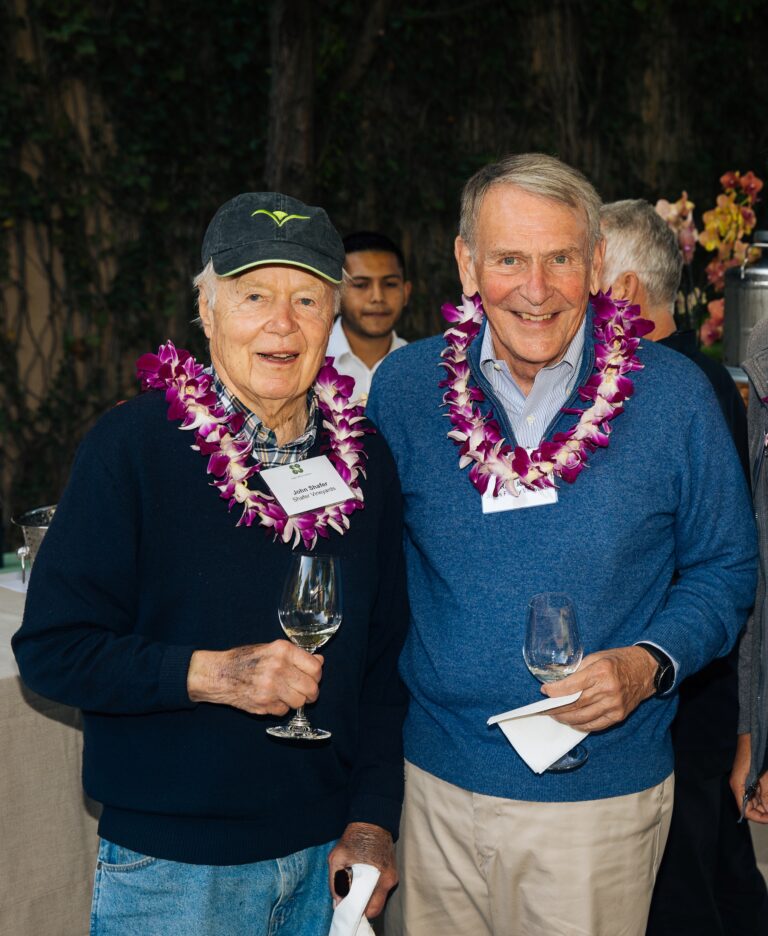 Two men stand next to one another wearing leis and smiling